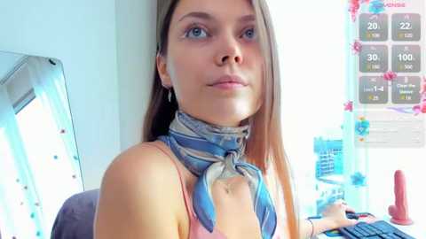 Video of a young woman with light skin, blue eyes, and long brown hair, wearing a blue scarf and a pink tank top, sitting at a desk with a computer and a red dildo on the right.