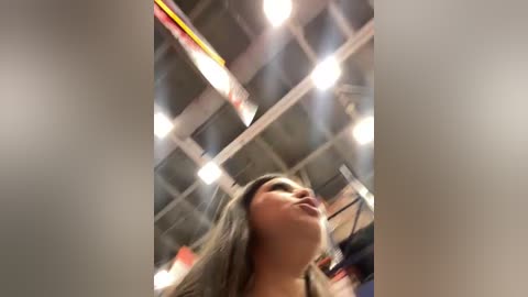 A video of a woman with light skin and long, wavy brown hair, smiling upwards, taken in a dimly lit room with a high ceiling and visible pipes, featuring a blurred background.