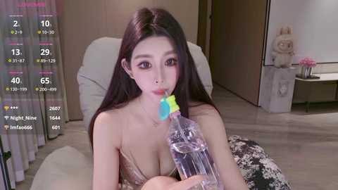 Video of an East Asian woman with long black hair, wearing a revealing gold top, drinking from a plastic bottle with a blue straw in a modern bedroom with a fluffy bed and a plush bunny.