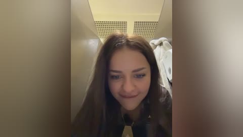 A video of a young woman with long, dark hair, wearing a dark jacket, leaning over the edge of a narrow, beige, utility closet.