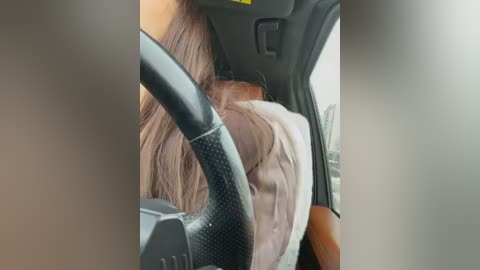 A video of a woman with long brown hair, wearing a light-colored shirt, driving a car with a black steering wheel and gray dashboard. The background shows the car's interior and a blurry outside view.
