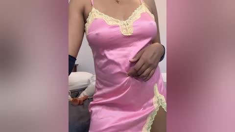 Video of a light-skinned woman with medium-sized breasts, wearing a pink satin nightgown with yellow lace trim, standing in a dimly lit room with blurred background.