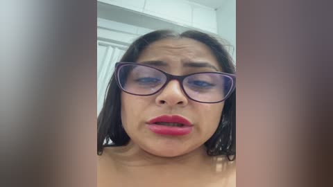 Video of a woman with medium skin tone, dark hair, wearing large purple-rimmed glasses, red lipstick, and no visible clothing, indoors with white walls and vertical blinds in the background.
