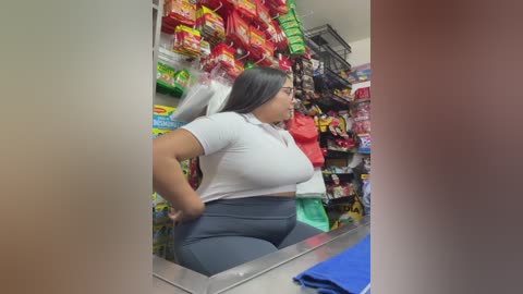 Video of a Latina woman with medium skin tone, long black hair, wearing a white polo shirt and high-waisted gray pants, working behind a cash register in a brightly lit convenience store with shelves filled with colorful snacks.