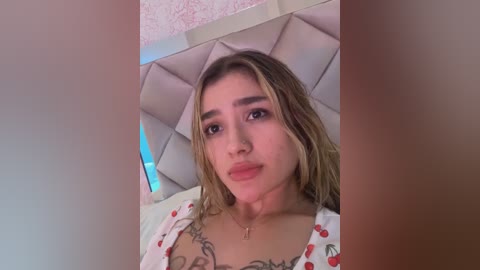 A video of a young woman with light skin, shoulder-length blonde hair, and a medium build, wearing a white top with cherry designs, lying on a bed with a pink quilt.