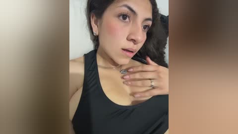 Video of a young woman with olive skin, dark hair, and a black tank top, wearing a silver ring on her left hand. She gazes upward, her face flushed. Background is blurred.