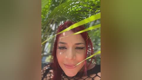 Video of a woman with fair skin, red hair, and dark eye makeup, partially obscured by green palm fronds. She's wearing a black fishnet top. The background is a lush, green outdoor setting.