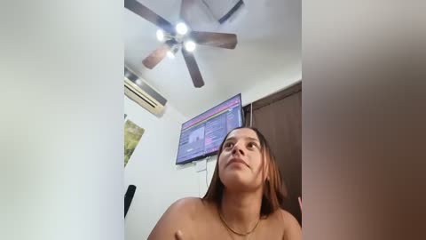 Video of a topless woman with medium brown skin and straight brown hair, standing in a small room with a ceiling fan, air conditioner, and TV on the wall.
