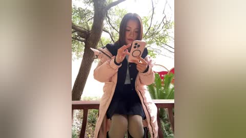 A video of an Asian woman with long black hair, wearing a pink puffer jacket, black dress, and beige tights, sitting on a wooden balcony, taking a selfie with a pink phone.