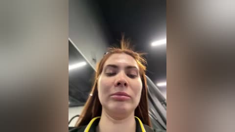 Video of a young Caucasian woman with long red hair and fair skin, wearing a black jacket, captured from a low angle in a dimly lit, metallic, indoor setting.