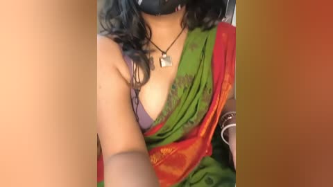 Video of a woman with medium brown skin wearing a green and red sari with a purple bra visible, black mask, and necklace, taken indoors.