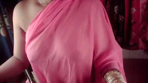 A video of a woman in a vibrant pink, draped saree with gold jewelry, standing against a backdrop of red and dark fabric, emphasizing traditional Indian attire.