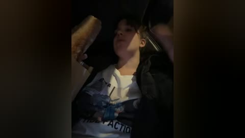 Video of a young boy, light-skinned with short dark hair, wearing a white t-shirt with a blue and black design, eating a large pastry inside a dimly lit car.