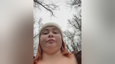 Video of a plus-size woman with fair skin, red hair, and a white knit hat, standing in a snowy forest with bare trees. She has a neutral expression.