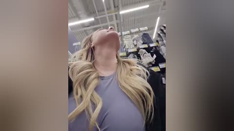 Video of a blonde woman with long, wavy hair, wearing a light blue shirt, looking upwards in a brightly lit, modern store with white ceiling and fluorescent lights.