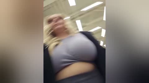 A blurry video of a blonde woman in a tight, light grey sports bra and black jacket, captured from a low angle in a brightly lit room with visible ceiling panels.