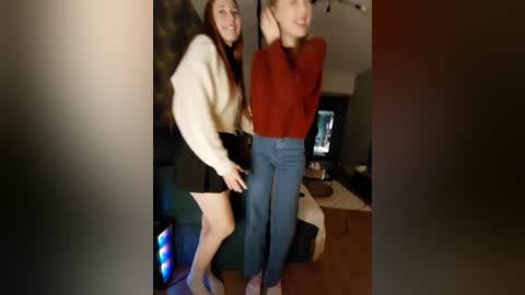 Video of two young women, one in a white sweater and black shorts, the other in a red sweater and blue jeans, playfully dancing in a dimly lit living room.