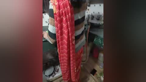 A video of a person wearing a striped shirt and a red sari with floral patterns, standing in a cluttered room with shelves and boxes in the background.