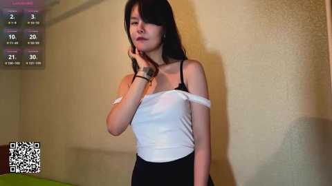 Video of a young Asian woman with straight black hair, wearing a white off-shoulder top and black pants, standing against a textured yellow wall. A digital clock and QR code are visible in the corner.