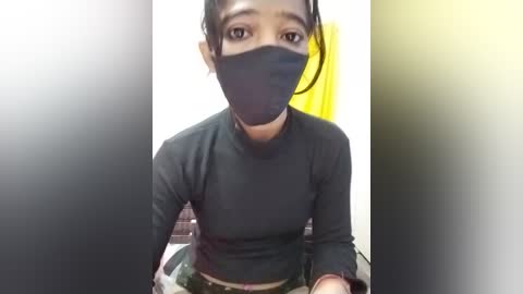 Video of a young woman with light brown skin, wearing a black face mask, black long-sleeved top, and dark hair tied back, seated in a brightly lit room with a yellow object in the background.