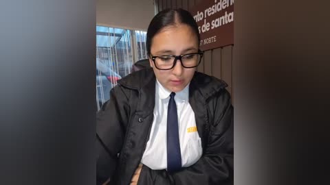 Video of a young woman with medium skin tone, dark hair, and black-rimmed glasses, dressed in a black blazer, white shirt, and navy tie, sitting in a government office with a bilingual sign and window blinds in the background.