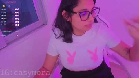 Video of a young woman with medium skin tone, wearing glasses, a white T-shirt with pink Playboy bunny logo, and black shorts, sitting in a dimly lit room with purple lighting.