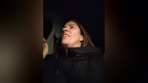 Video of a smiling, middle-aged woman with shoulder-length brown hair, wearing a black jacket, holding a small, dark object, set against a dimly lit, out-of-focus background.