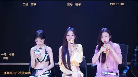 Video of three East Asian women, one in a black bikini, one in a yellow blouse, and one in a pink blouse, seated on black chairs, singing into microphones on a dark stage.