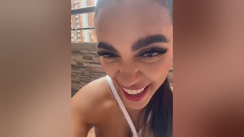 Video of a woman with light brown skin, dark hair, and black eyeliner, smiling and looking directly at the camera. She wears a white strapless top. Background features a stone wall and metal railing.