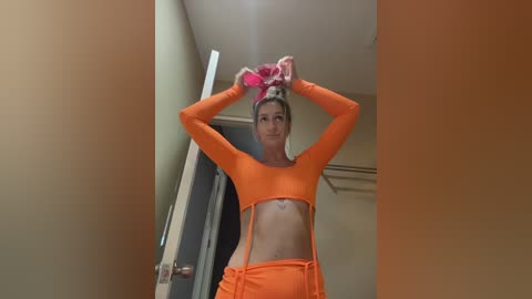 A video of a slender, fair-skinned woman with a toned physique wearing an orange crop top and matching shorts. She has a tattoo on her stomach and is adjusting a pink headband in a beige hallway.
