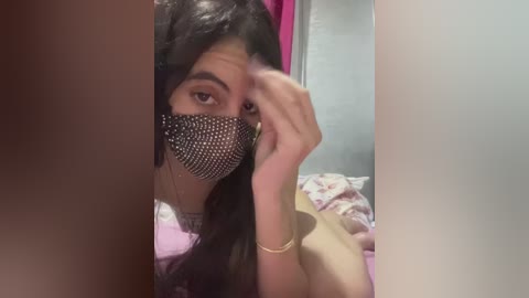 A video of a young woman with long dark hair, wearing a black sequined mask, lying on a bed with pink sheets, partially obscured by a large white object.