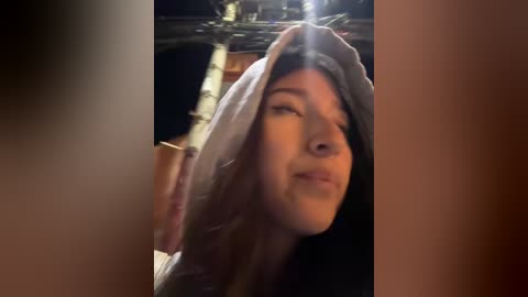 Video of a young woman with light brown hair, wearing a hooded robe, looking upwards with a contemplative expression. Background features a dimly lit, possibly religious setting with wooden beams.