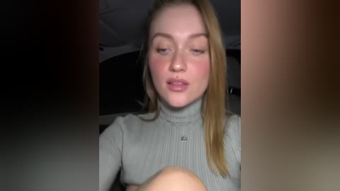 A video of a young, fair-skinned woman with straight blonde hair, wearing a grey ribbed turtleneck sweater, sitting in a car, her lips slightly parted.