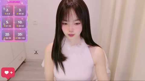 Video of a young East Asian woman with long black hair and fair skin, wearing a sleeveless white top, sitting in a minimalist room. Background includes a white wall and a purple virtual rating display.