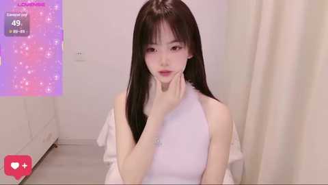A video of an Asian woman with long black hair, wearing a white sleeveless dress, sitting on a white couch in a minimalist room. A digital overlay shows a social media post with 45 likes.