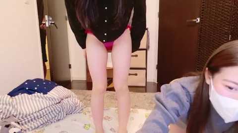 A video of a woman in a bedroom, wearing a black button-up shirt and red panties, standing with legs apart, facing away. Another woman in a mask, blue shirt, and gray pants is in the foreground.