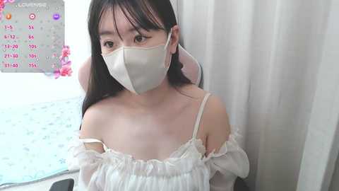 Video of an Asian woman with long black hair, wearing a white off-shoulder dress and face mask, standing indoors near a window with floral curtains.