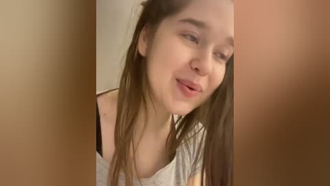 Video of a young woman with long, straight brown hair, fair skin, and a light gray off-the-shoulder top, smiling slightly. Background shows a beige wall.