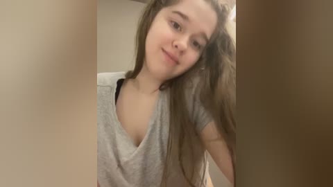 A video of a young Caucasian woman with long brown hair and fair skin, wearing a gray V-neck shirt, smiling slightly. The background is a plain beige wall.