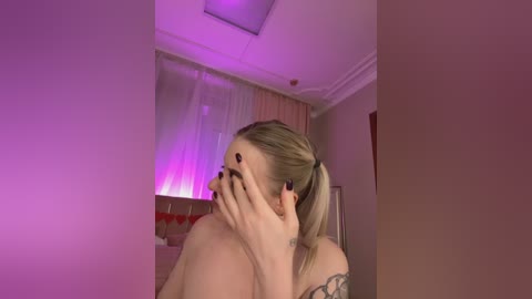 Media: Video of a topless blonde woman with a ponytail, covering her face with her hands, standing in a dimly lit room with purple lighting.