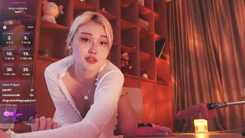Video of a young Asian woman with light skin and blonde hair, wearing a white cardigan, leaning on a wooden table, surrounded by shelves with stuffed animals, dim lighting, and a microphone.