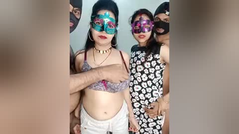 Video of three women with dark skin, wearing colorful, ornate masquerade masks, revealing their faces partially. They are dressed in vibrant outfits, with one in a floral dress and another in a patterned top.