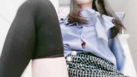 A video of a young woman with light skin and long dark hair, wearing a light blue blouse with a ruffled collar and black thigh-high socks, sitting on a white bed with a black and white checkered skirt.