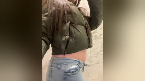 Video of a woman with light skin and long dark hair, wearing a green puffy jacket, unzipped to reveal her midriff, and light blue jeans. Background features a marbled wall.