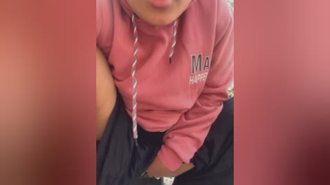 A video of a woman in a pink hoodie with \"MDA\" text and a black skirt, leaning over with a silver necklace dangling, in a red bathroom stall.