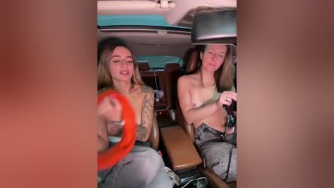 Video of two topless women with small breasts in a car, wearing jeans; one with orange hair, the other with long brown hair, smiling, car interior visible.