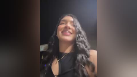 Video of a joyful, smiling woman with long, wavy black hair, wearing a black sleeveless top and a gold necklace, set against a dark background with blurred edges.