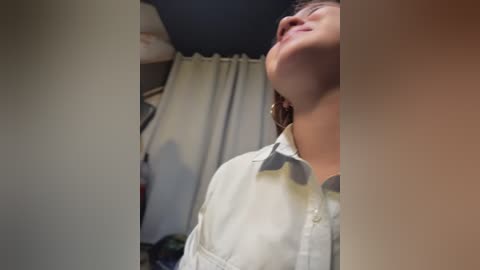 A video of a woman with short brown hair in a white shirt, standing in a dimly lit room with beige curtains and a white lampshade.