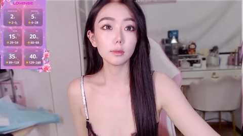 Video of a young Asian woman with long black hair, wearing a black lace bra, in a well-lit bedroom with a makeup vanity.