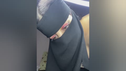 A video of a person with short, dark hair wearing a black blindfold, partially obscuring their eyes. The background shows a dimly lit room with a beige wall and a yellow pillow.